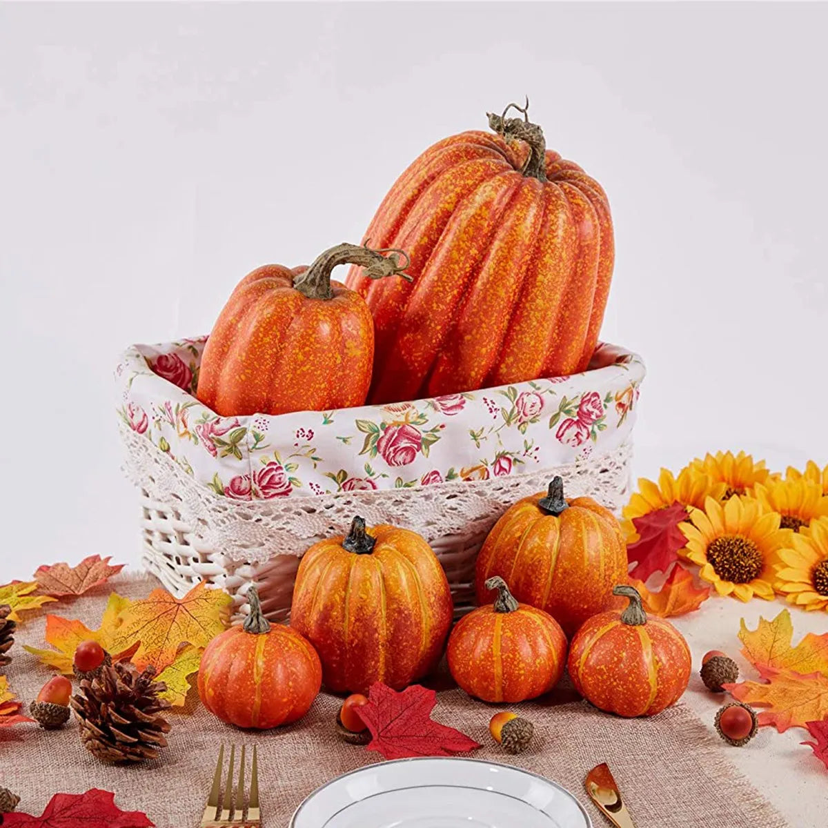 A set of seven orange artificial pumpkins arranged in a woven basket on a table with autumn leaves and flowers.