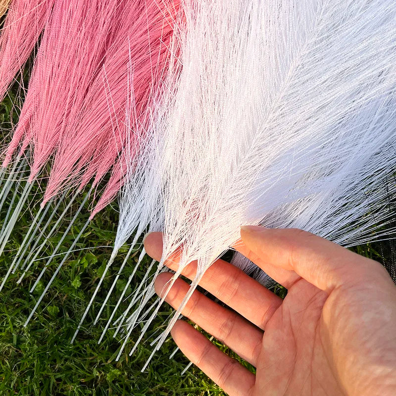 Primer plano de penachos artificiales de pampas rosadas y blancas sostenidos en una mano contra hierba verde.