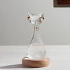 A clear glass bottle shaped like an Egyptian cat head filled with water, resting on a wooden base.