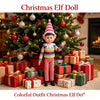 A Christmas elf doll dressed in colorful striped clothing standing in front of a decorated Christmas tree and wrapped gifts.