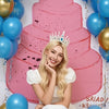 A blonde woman in a white dress poses in front of a multi-tiered pink cake backdrop decorated with gold and blue balloons.