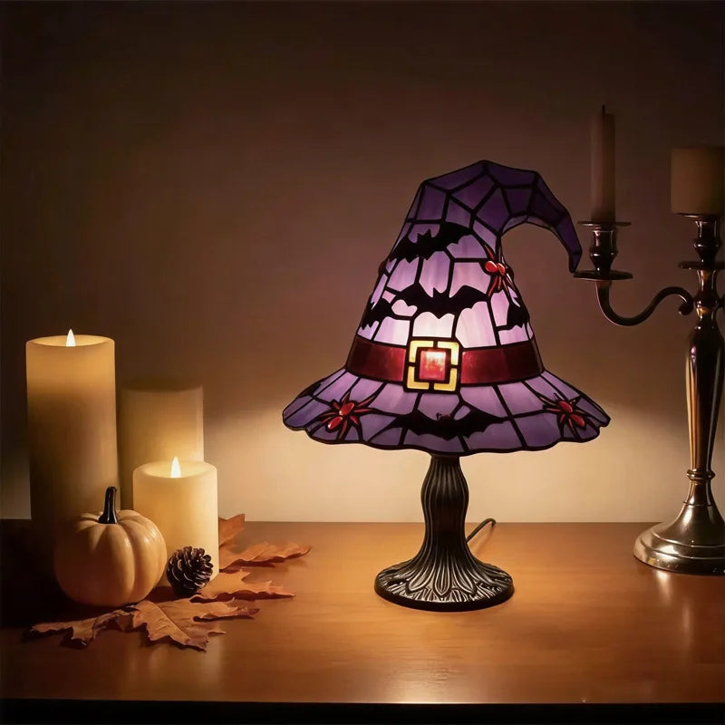 A purple witch hat night light with a stained glass effect sits on a wooden table next to several candles and autumn leaves.
