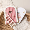 A pink wooden fairy elf door miniature decoration with a heart cutout and a small tooth box placed on a woven basket.