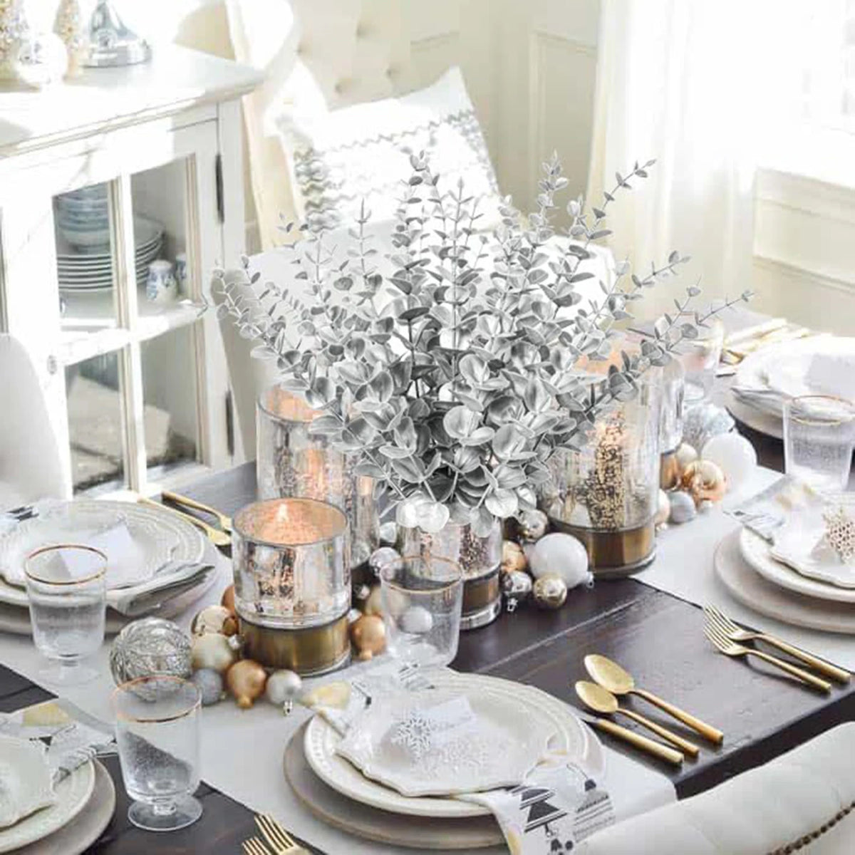 A bouquet of silvery-green artificial eucalyptus leaves displayed in a glass vase on a dark wooden table.