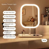 A white square tricolor light vanity mirror with a round LED frame is displayed on a bedside table.
