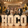 Five young adults posing in front of large marquee light-up letters spelling H O C O for a party decor event.