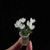 A hand holds a small glass vase filled with white handcrafted mini flowers against a black background.
