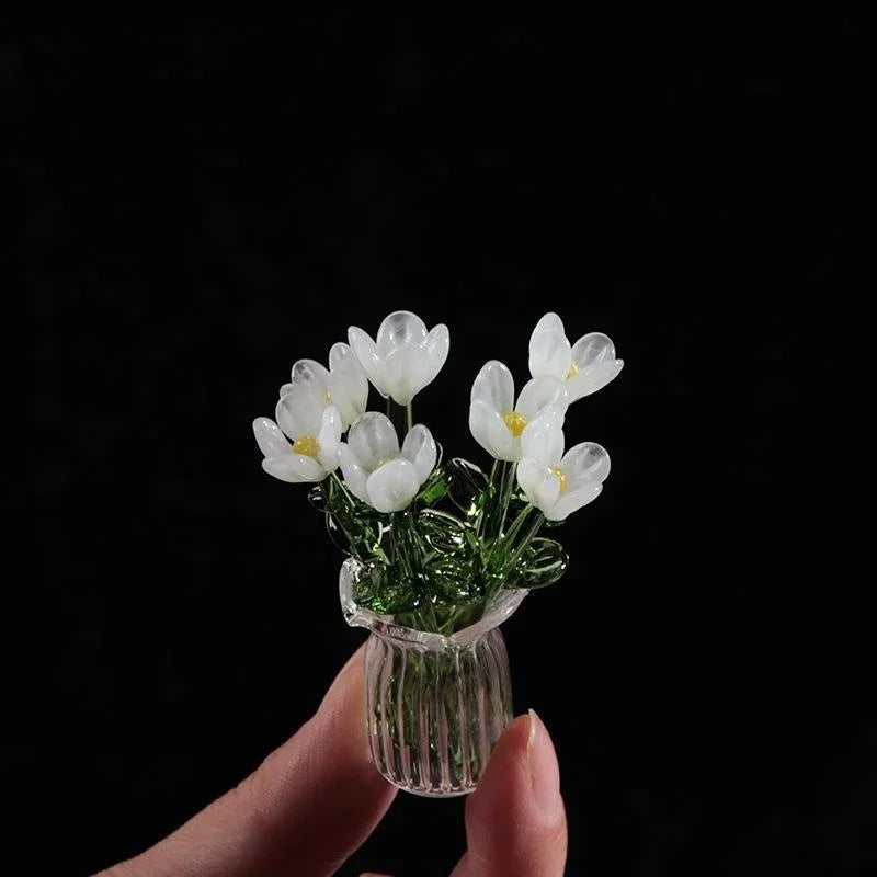 A hand holds a small glass vase filled with white handcrafted mini flowers against a black background.