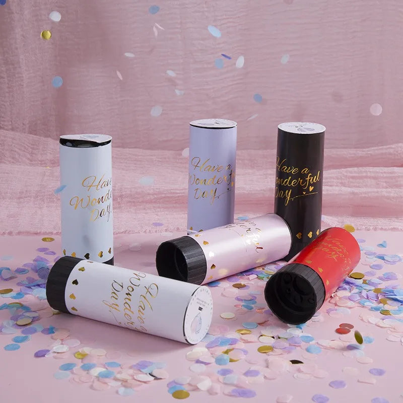 A collection of hand twist confetti cannons in various colors, including silver, purple, and black, scattered on pink confett