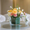 A bouquet of yellow, white, and pink roses and greenery in a dark green square planter.