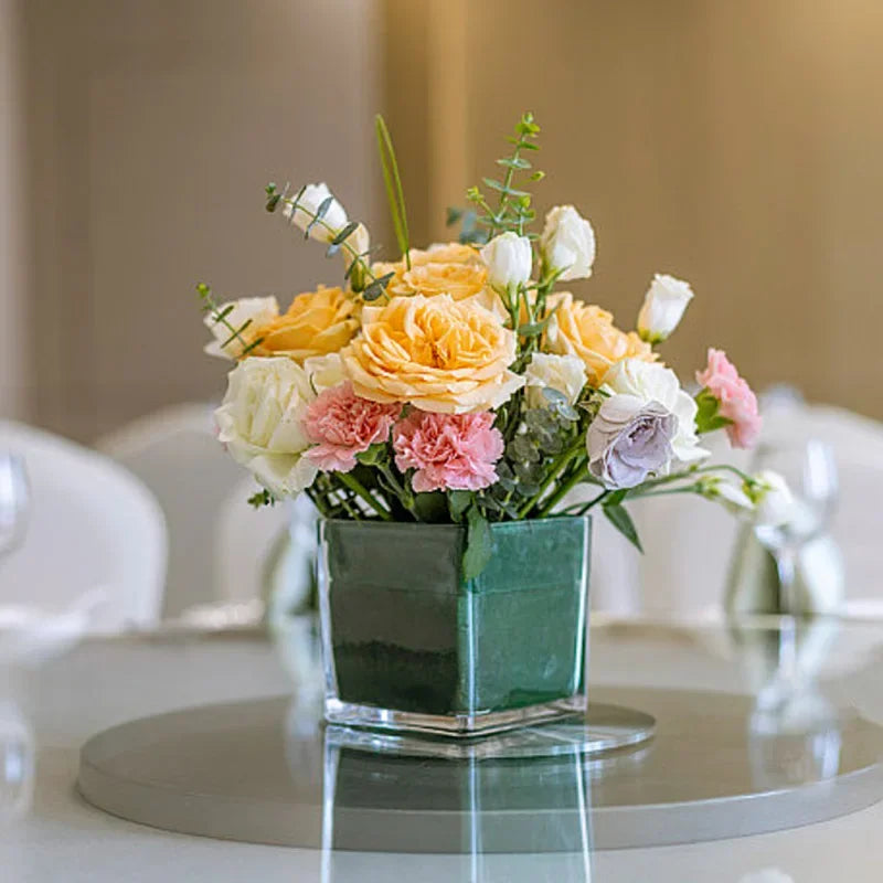 A bouquet of yellow, white, and pink roses and greenery in a dark green square planter.