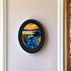 An abstract oval painting featuring vibrant orange, yellow, and blue tones set in a black frame.