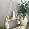 A handmade macrame wood wall shelf with tassels hanging on a gray wall, surrounded by green plants.