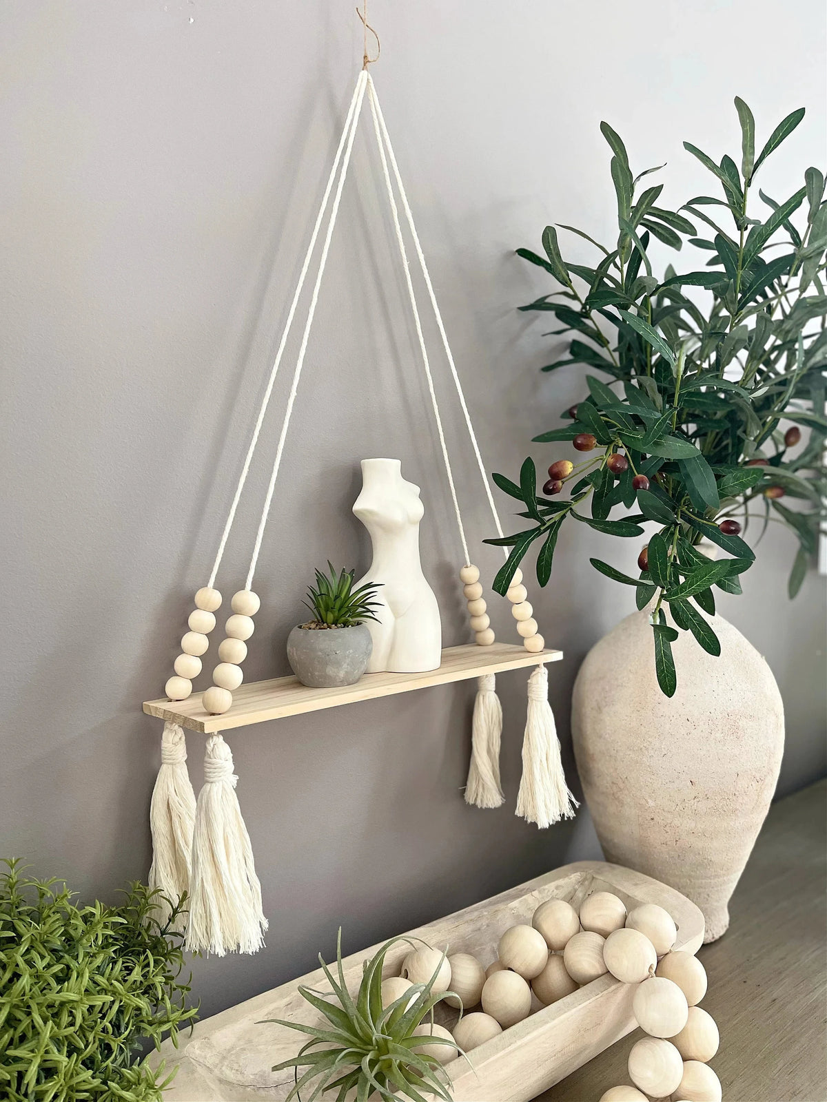 A handmade macrame wood wall shelf with tassels hanging on a gray wall, surrounded by green plants.