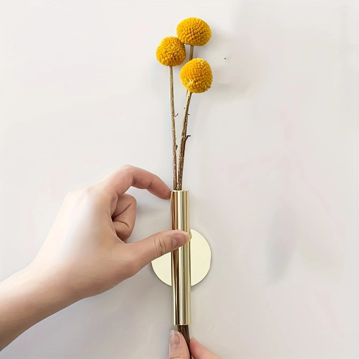 Eine Hand hält eine goldfarbene, wandmontierte Blumenvase mit getrockneten gelben Blumen vor weißem Hintergrund.