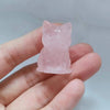 Un ornement de chat en cristal rose sculpté à la main est tenu dans la main d'une personne sur un fond gris clair.