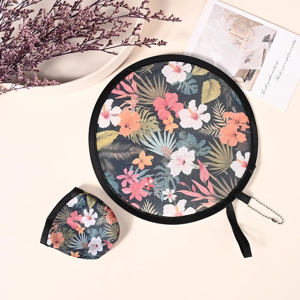 A black fabric portable foldable cooling handheld fan with a floral and tropical leaf pattern is shown on a light surface.