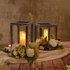 A rustic wooden candle holder lantern with a glowing candle and floral arrangement on a wooden surface.