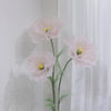 A large artificial silk pink poppy flower with a green stand displayed against a white wall.