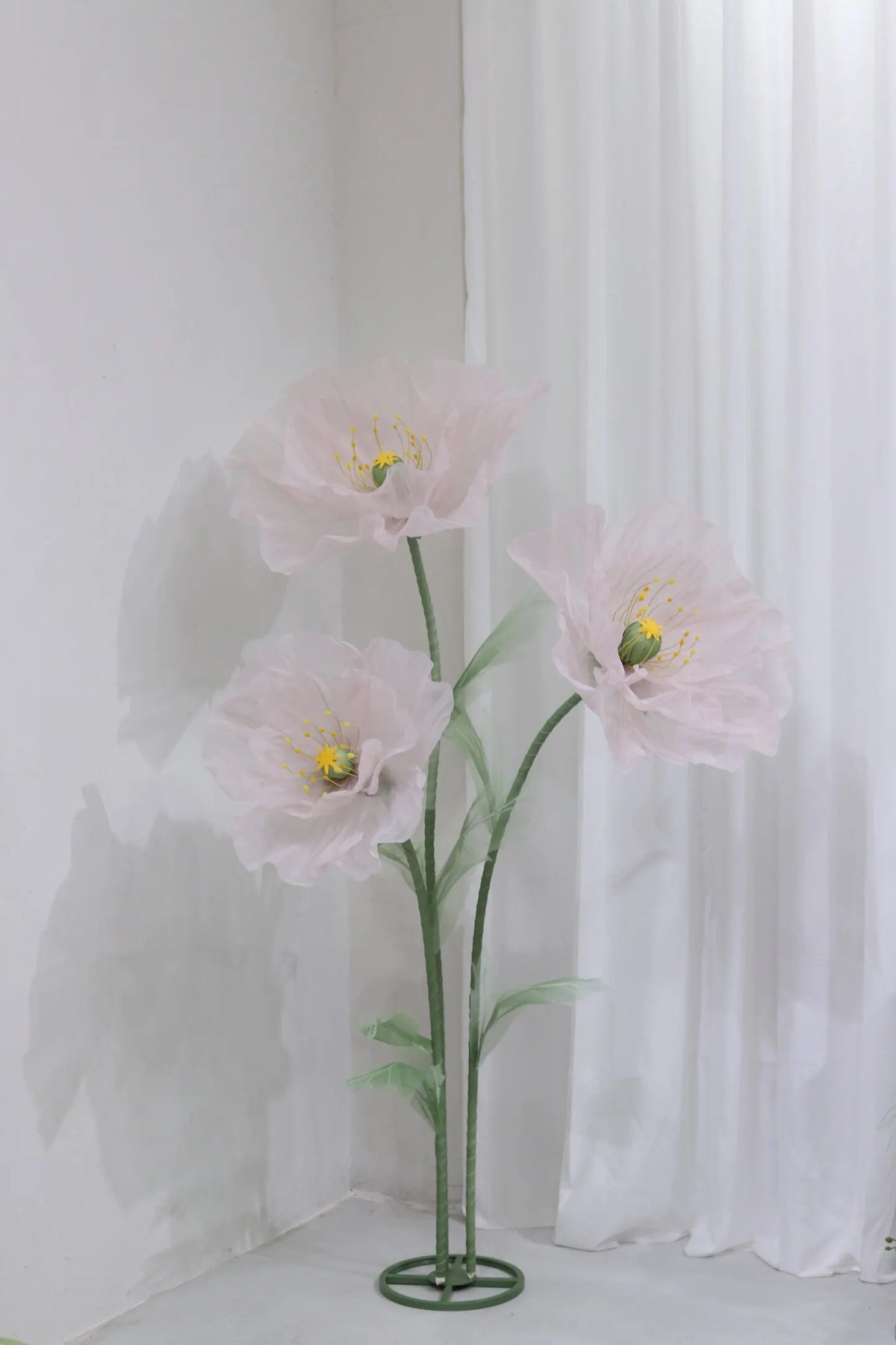 A large artificial silk pink poppy flower with a green stand displayed against a white wall.
