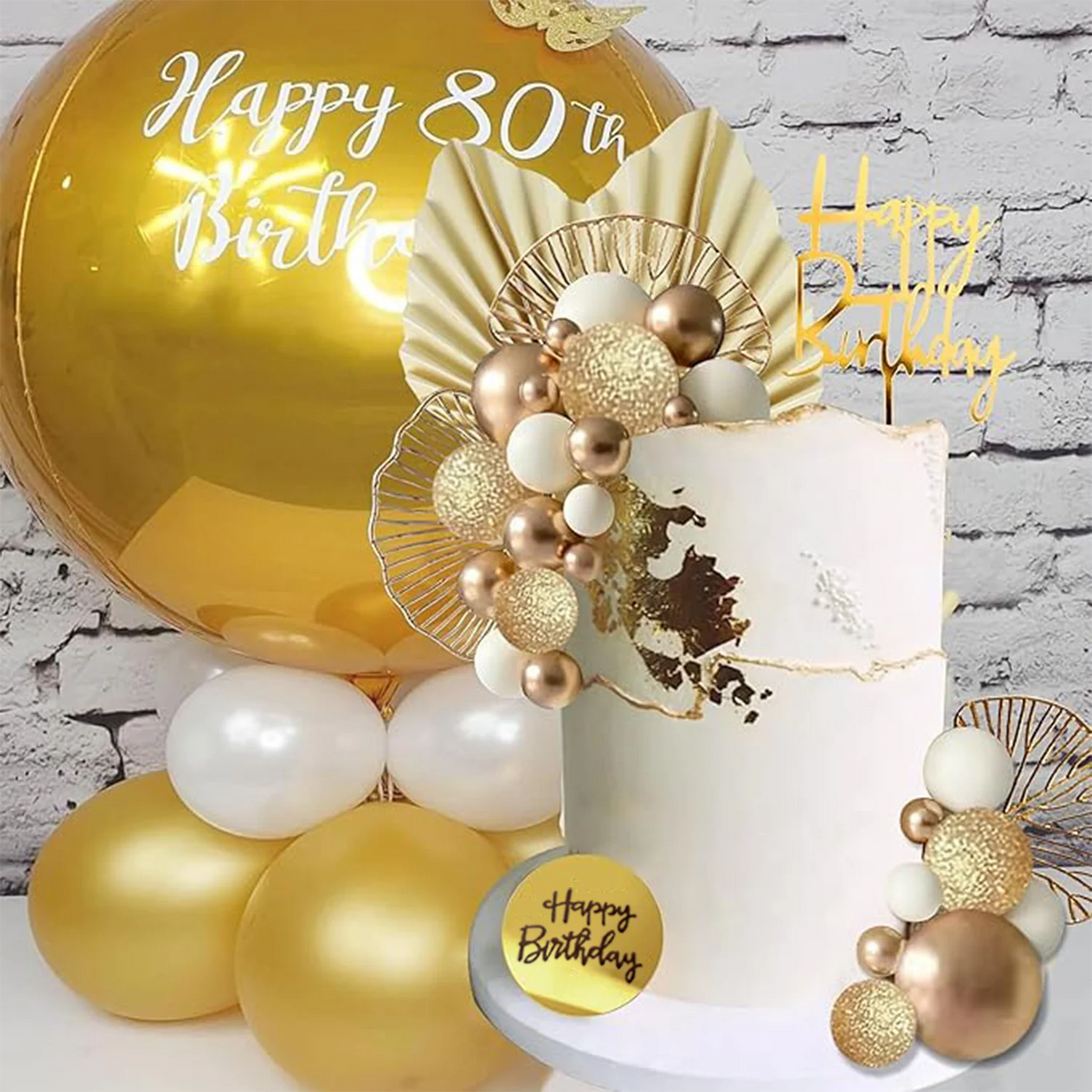 A white cake decorated with gold leaf and pearl accents, surrounded by gold and white balloons.