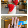 A hand holds a bright red strawberry charm and keychain against a blurred background of a modern cafe.