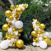 A balloon arch kit featuring gold and white balloons set up outdoors against a green hedge and fence.