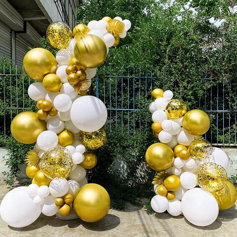 A balloon arch kit featuring gold and white balloons set up outdoors against a green hedge and fence.