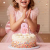 A young girl holds her hands over a pink glitter number cake decorated with a bow and lit candles.