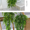 Plante artificielle en rotin vert luxuriant, suspendue dans un pot blanc sur fond clair.