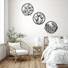 A set of three black line-art metal flower wall art pieces displayed on a white wall above a wooden floor and a light wood ch