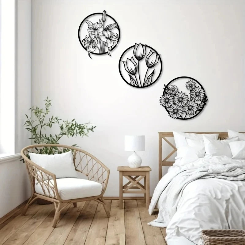 A set of three black line-art metal flower wall art pieces displayed on a white wall above a wooden floor and a light wood ch