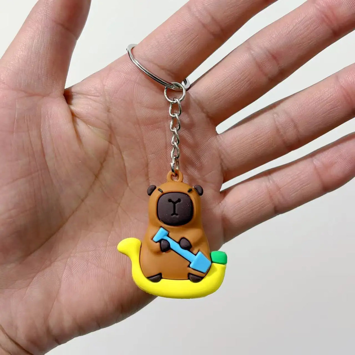 Un petit pendentif de capybara brun avec une pelle bleue accroché à un anneau argenté, tenu dans une main.