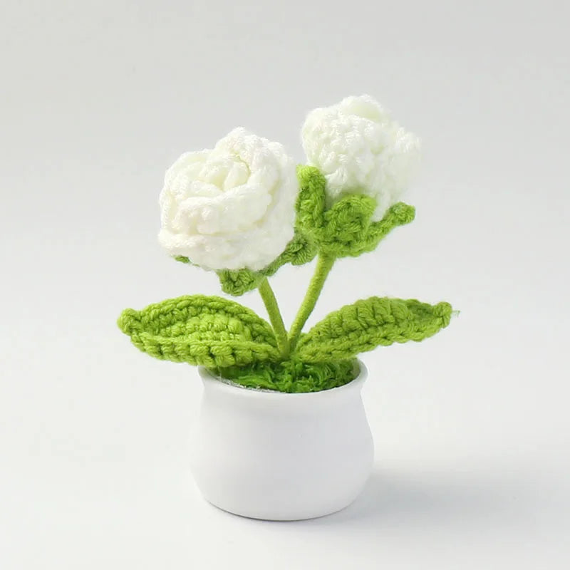 A hand-woven crochet white rose plant in a white pot with green leaves.
