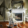A skeletal figure draped in black gauze fabric sits in front of a fireplace mantel decorated with spooky items.