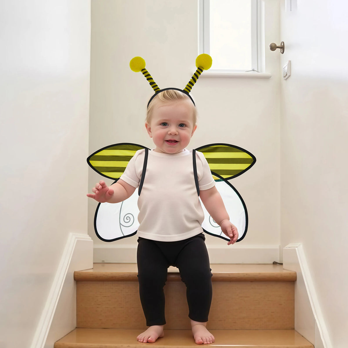 A toddler wearing an insect wing and headband costume set with yellow antennae and black wings standing on a wooden staircase