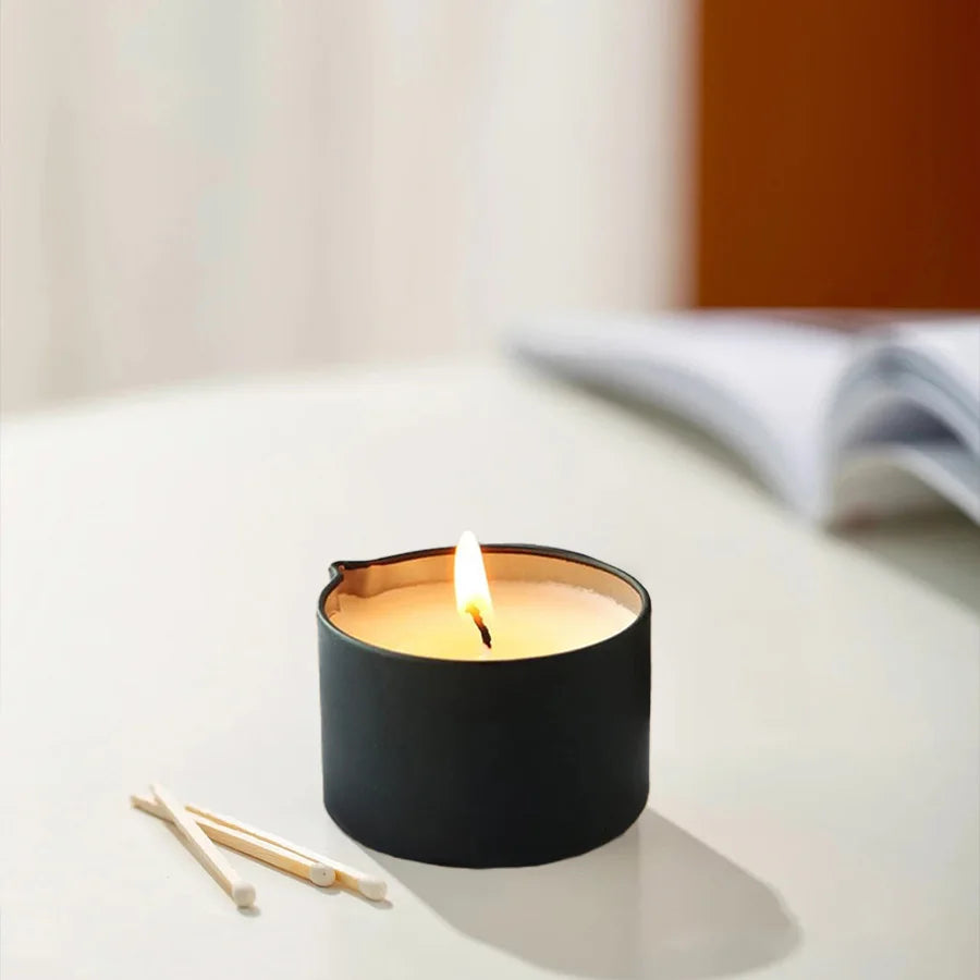 A black candle in a holder with a lit wick sits on a white surface next to wooden sticks.