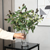 A hand holds a glass vase filled with an artificial olive tree branch featuring green leaves and dark berries.