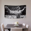A black and white tapestry featuring a muscular man with raised arms and the word CONQUER on a wall.
