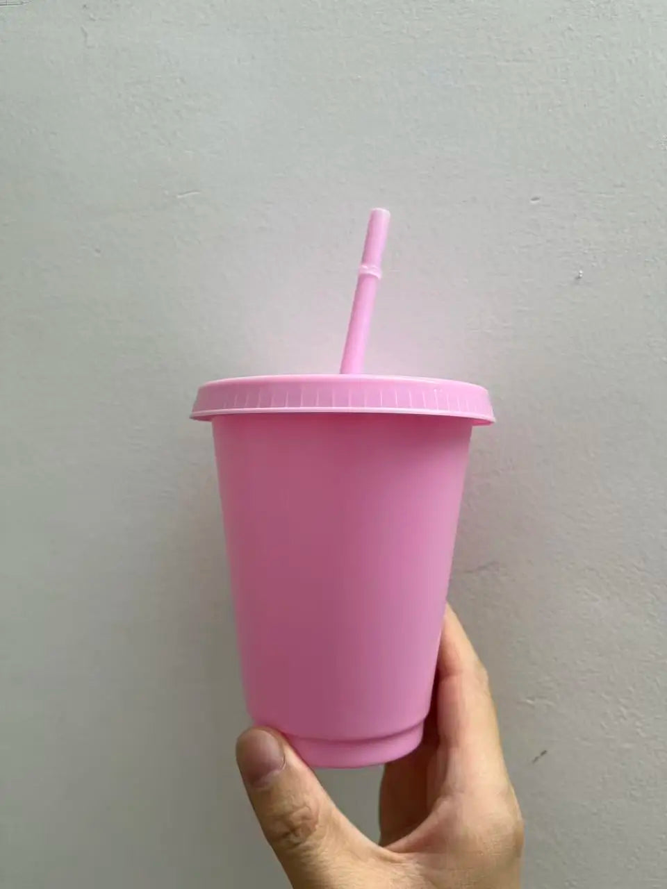 A hand holds a pink plastic cold cup with a lid and straw against a light gray wall.