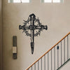 Black metal cross wall art with thorn details mounted on a beige wall above a wooden railing.