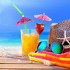 A glass of orange juice sits next to a strawberry and colorful mini paper umbrellas on a wooden beach surface.