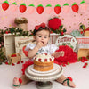 A baby is holding a strawberry cake on a stand in front of a pink backdrop decorated with strawberry motifs.