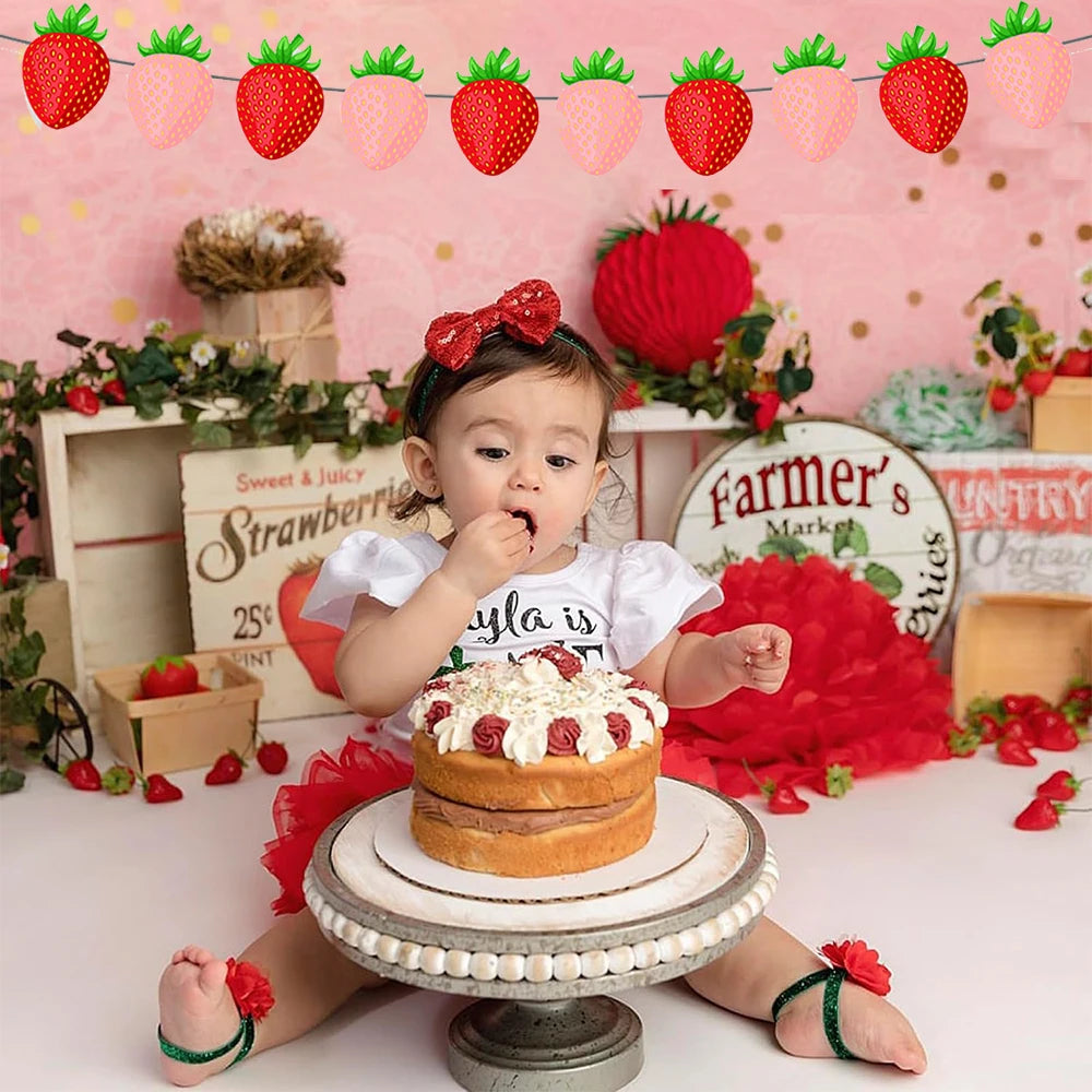 A baby is holding a strawberry cake on a stand in front of a pink backdrop decorated with strawberry motifs.