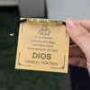 A close-up of a gold-toned stainless steel bracelet with an engraved tag featuring the text Dios Tiene El Control