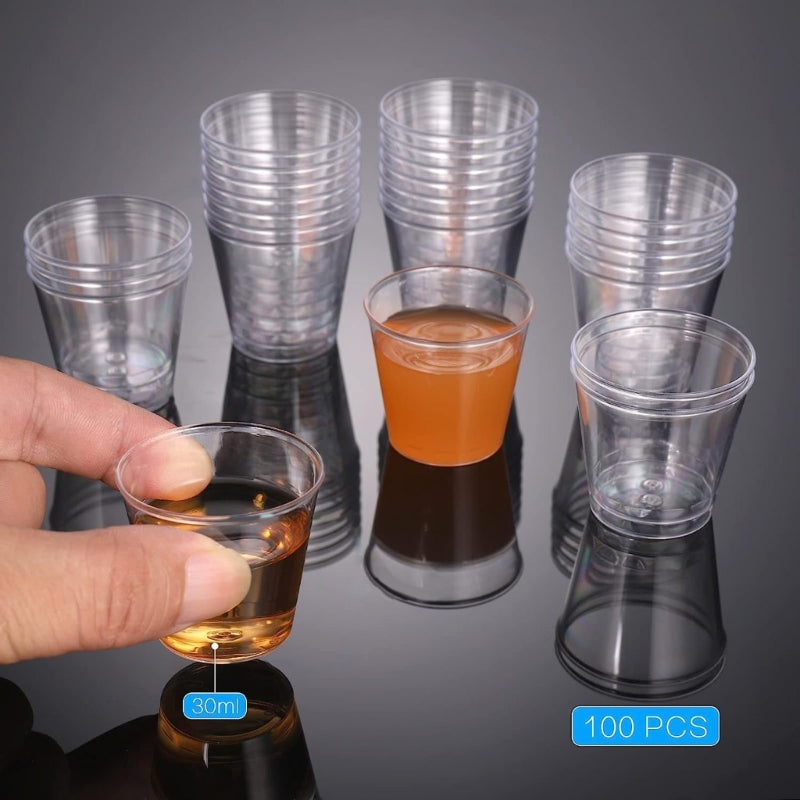 A hand holds a small glass of amber liquid next to a set of 100 clear reusable plastic shot glasses.