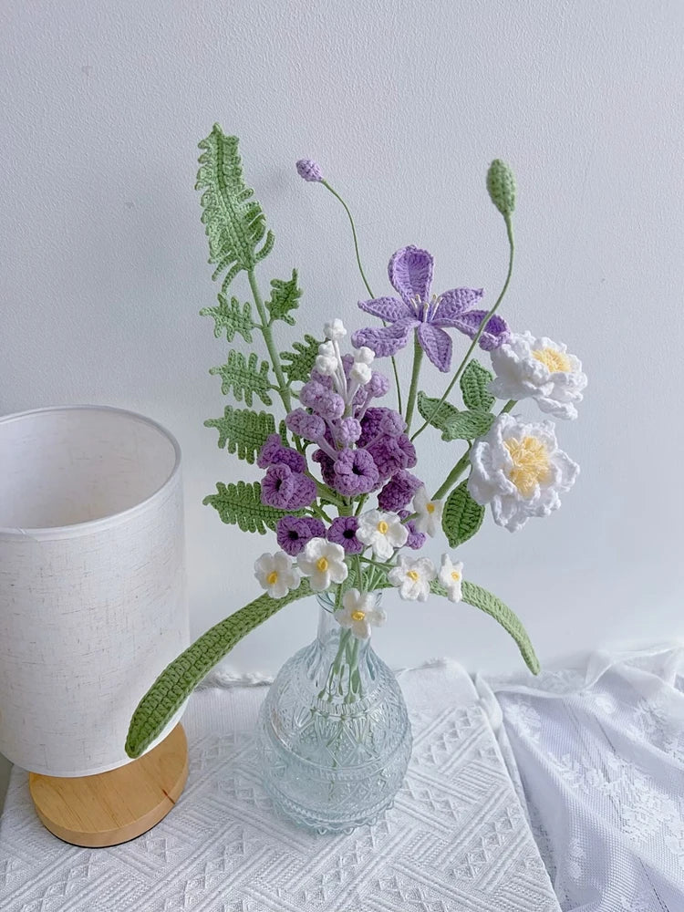 A handmade crochet flower bouquet featuring purple, white, and green flowers in a clear glass vase on a white surface.