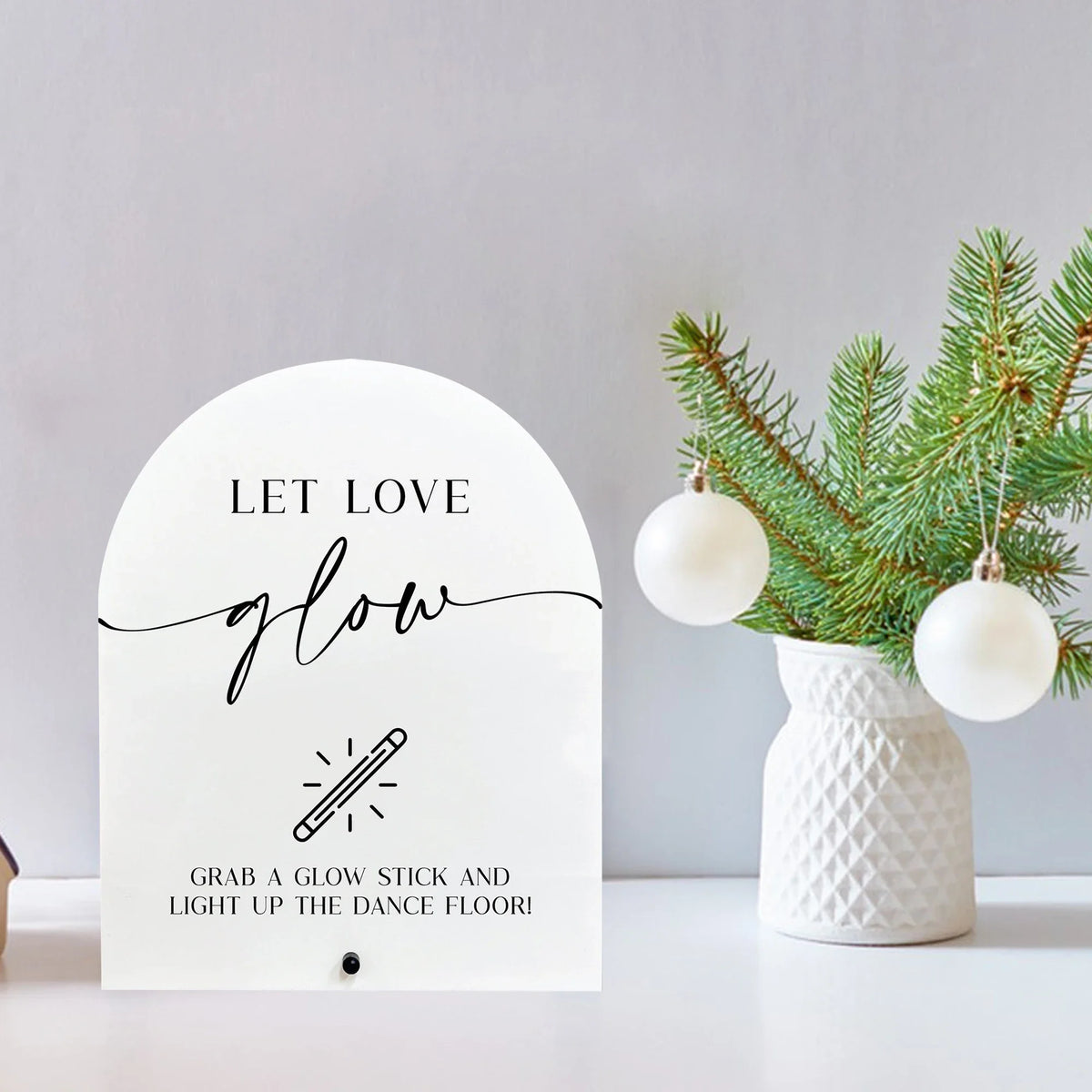 A white acrylic sign with black script that reads Let Love Glow and features a graphic of a glow stick.