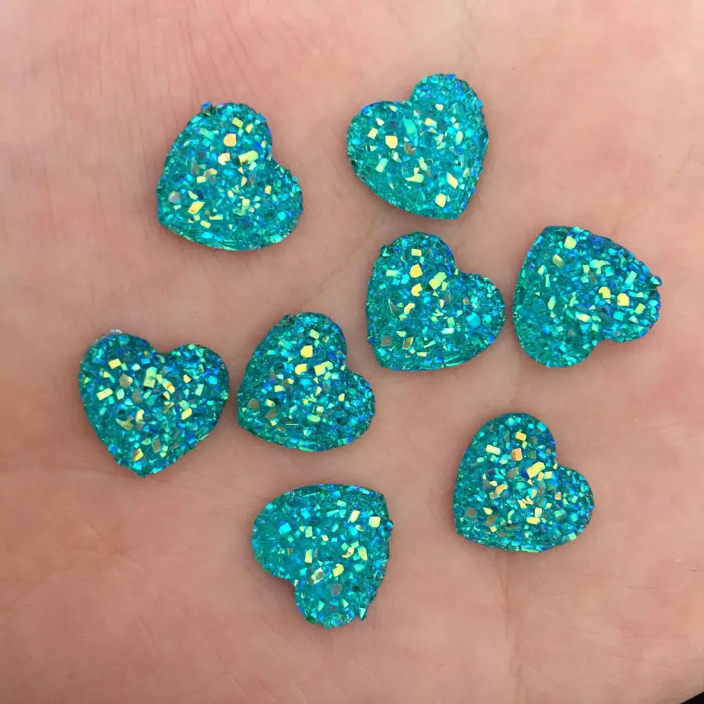 Eine Hand hält eine Sammlung kleiner, herzförmiger blaugrüner Strass-Verzierungen, die mit Glitzer funkeln.