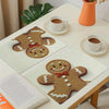 A set of four gingerbread man printed placemats on a white table with a cup and orange pot.